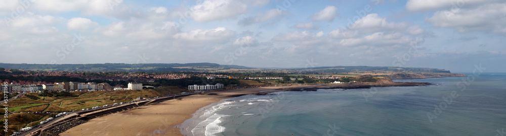 wide aerial panoramic view of scarborough north bay with buildings and beach huts behind the beach and a blue sunlit sea