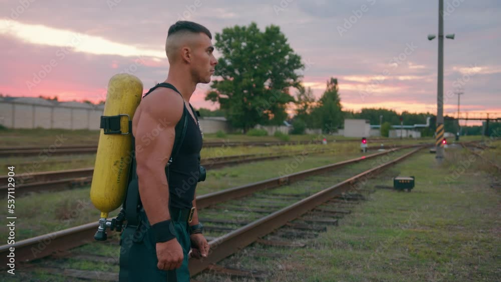 A young bodybuilder with a compressed air tank dragger on his back stands on the rails at sunset. Slow motion