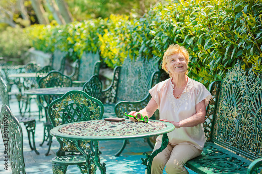 Obraz premium Mature attractive woman traveler sitting alone on the terrace of coffee shop in beautiful park in Bulgaria. Active life of the elderly in retirement, active seniors
