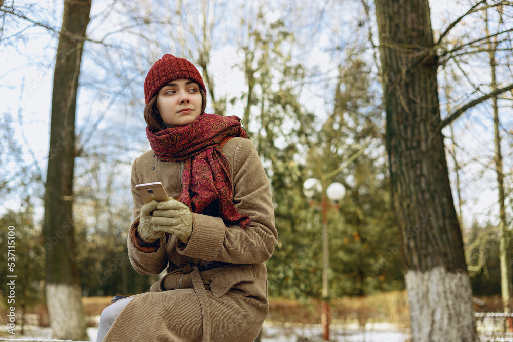 Low angle of thoughtful young woman in winter outerwear browsing mobile phone and looking away while chilling in snowy park 