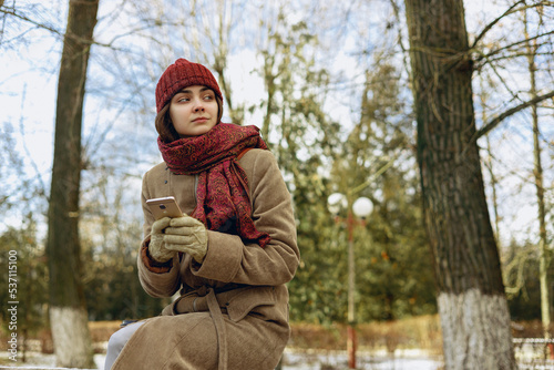 Low angle of thoughtful young woman in winter outerwear browsing mobile phone and looking away while chilling in snowy park  © Dzmitry