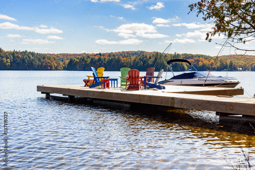 Fototapeta premium Motorboat tied up to a wooden pier on a lake on a sunny autumn day. Colourful armchairs are on the pier.