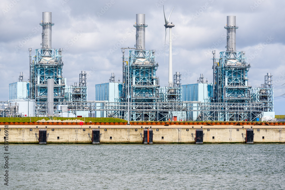 Fototapeta premium Gas fired power station on a harbor on a cloudy summer day. A wind turbine is in background. Eemshaven, Netherlands.