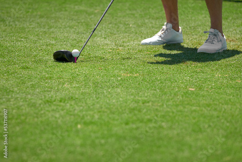 Detail of golf club about to hit ball by sneakers on green grass