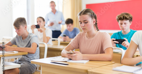 Concentrated teenage girl with group of fellow students sitting at lesson in classroom, using mobile phone