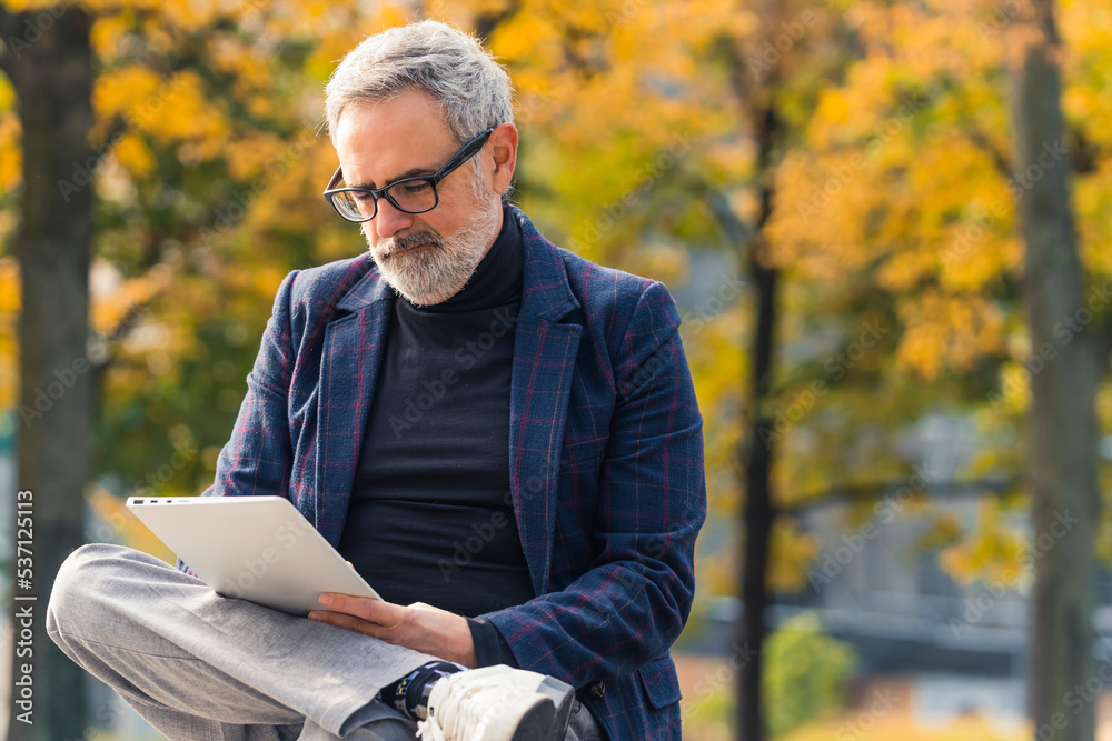 Fototapeta premium Elegant retired caucasian man with eyeglasses and gray hair and gray facial hair reading a book on his laptop while sitting on bench at park. Medium outdoor shot. Autumn colors. High quality photo