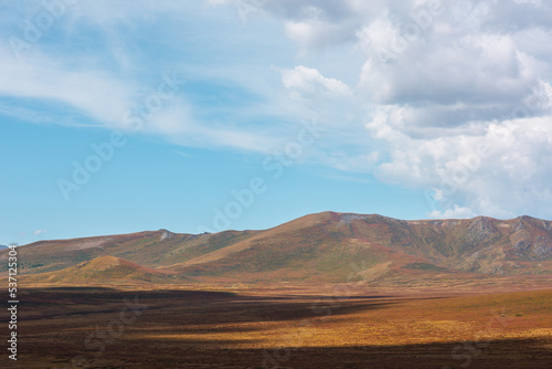 Motley autumn landscape with sunlit high mountain plateau and mountain range under dramatic cloudy sky. Vivid autumn colors in mountains. Sunlight and beautiful shadows of clouds in changeable weather