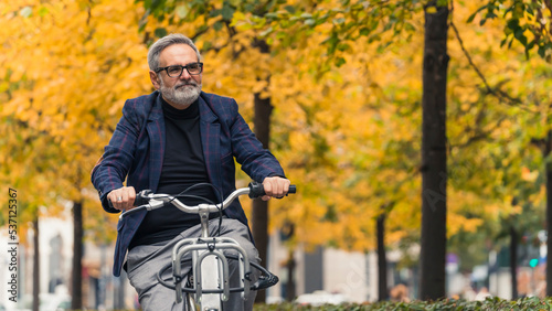 Happy senior actively spending time outdoors. Autumn in the city. Happy caucasian man in warm blazer riding his bike in nearby park. Blurred yellow-leaf trees in the background. High quality photo