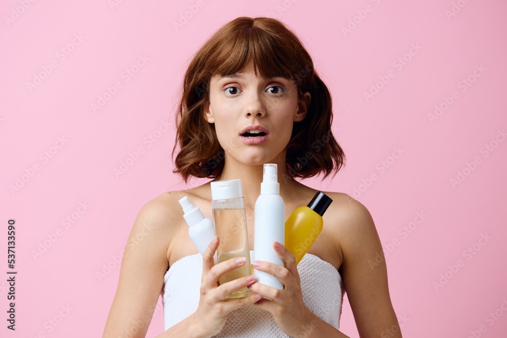 a young, pleasantly surprised woman stands on a pink background holding different jars of care cosmetics in her hands and happily looks into the camera with her mouth wide open. a young, pleasantly surprised woman stands on a pink background holding different jars of care cosmetics in her hands and happily looks into the camera with her mouth wide open.