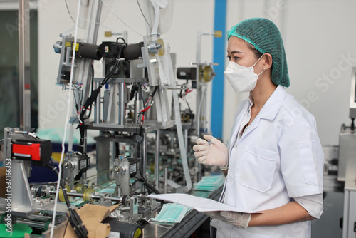 Worker woman in personal protective equipment or PPE inspecting quality of mask and medical face mask production line in factory, manufacturing industry and factory concept.
