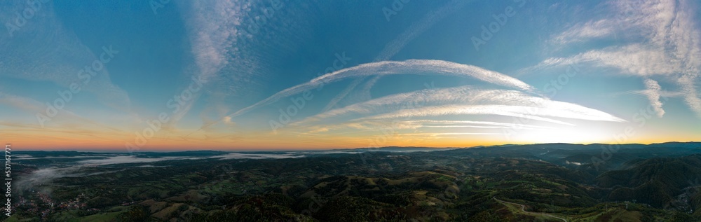 Aerial panorama view of a foggy sunrise with visible airplane contrails in a hilly area near Resita city, Romania. Captured with a drone