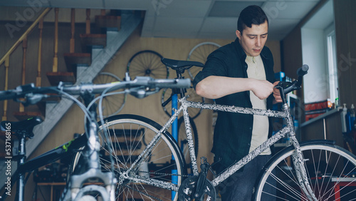 Experienced male master is checking and repairing bicycle handle-bar with special tools while working in workshop with numerous spare parts and equipment.