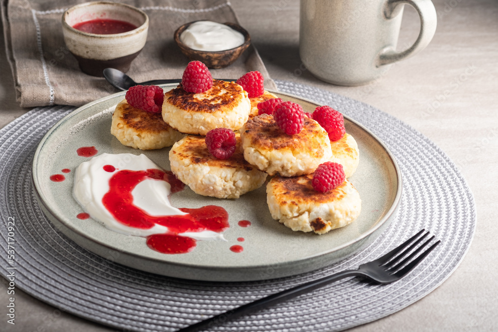 Cheesecakes or syrniki on a round plate with raspberry, sour cream and raspberry syrup.Healthy breakfast. Delicious dessert. Cheesecakes or syrniki on a round plate with raspberry, sour cream and raspberry syrup.Healthy breakfast. Delicious dessert.