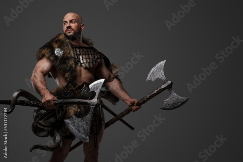 Studio shot of handsome ancient viking dressed in armor and fur holding two axes.