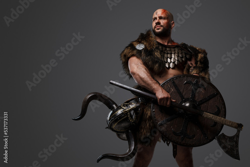 Studio shot of barbaric viking with bloody face holding shield and axe isolated on grey.