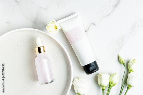 top view of cosmetics set - beauty face oil in dropper bottle and unbranded tube with cream or mask on marbal table with eustoma flowers. Trendy flat lay stillife photo