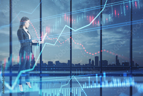 businesswoman with laptop looking at glowing candlestick forex chart hologram on blurry panoramic office window frame with blurry night city view. Stock, investment, market and money concept. 
