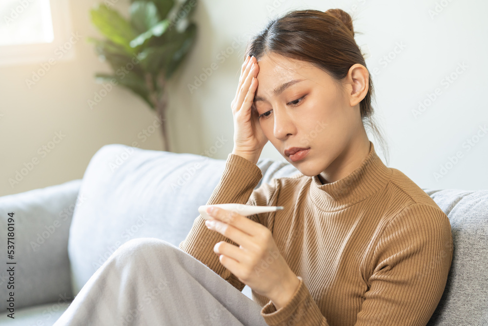 Sick, influenza asian young woman, girl headache have fever, hand touching forehead, holding thermometer for check measure body temperature, illness while sitting rest on sofa at home. Health care. Sick, influenza asian young woman, girl headache have fever, hand touching forehead, holding thermometer for check measure body temperature, illness while sitting rest on sofa at home. Health care.