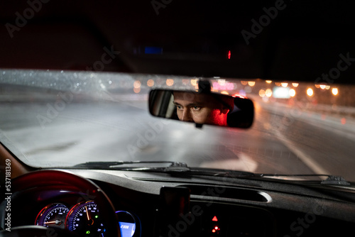 Look of man is reflected in rear-view mirror traveling in car in evening hurries home after work. Speedometer with neon lights and eyes young guy working as driver or cabbie and enjoying city at night