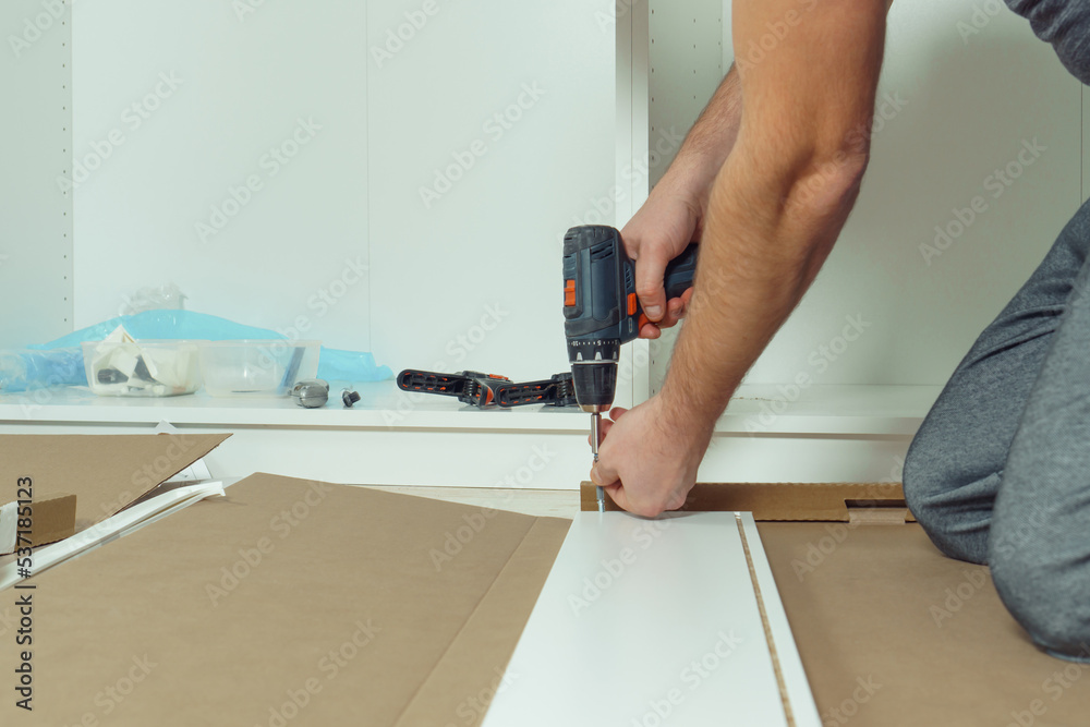 Male hands hold cordless electric screwdriver, screw to fix walls of white drawer, assembling dresser or wardrobe on floor. Self-assembly of furniture at home, DIY, furniture repair and rearrangement