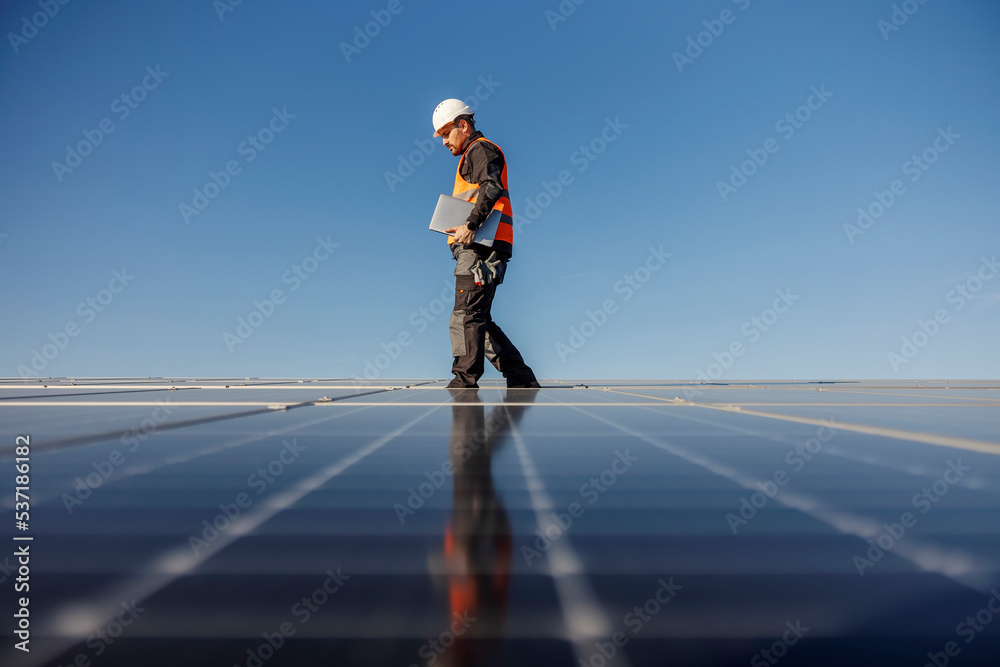 A supervisor is walking on the rooftop with solar panels and holding tablet under armpit. A supervisor is walking on the rooftop with solar panels and holding tablet under armpit.