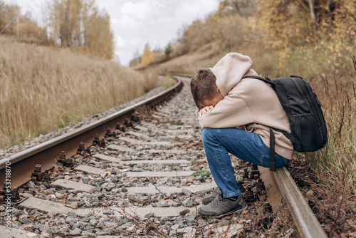 Sad teenager boy with backpack sitting on rails of railway. Mental health concept.