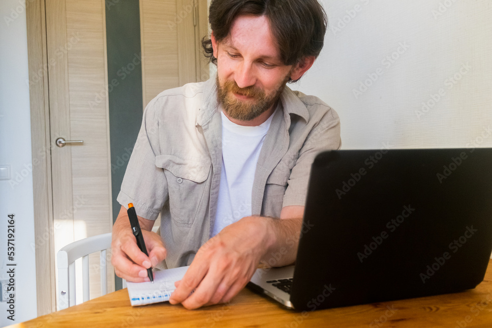 Bearded man smiling writes something in a notebook next to a laptop Bearded man smiling writes something in a notebook next to a laptop