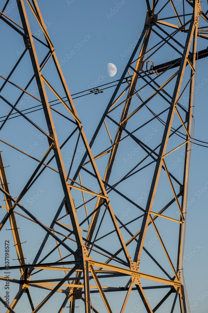 Detail of a tower of high voltage cables and the moon in the background in a clear sky