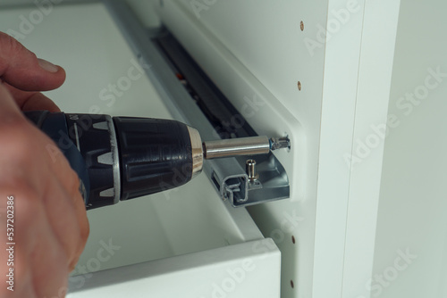Принти на полотні Male hands hold cordless electric screwdriver, screw to fix rail, assembling drawer of white dresser or wardrobe