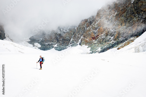 Mountaineer alpinist standing glacier ice snow covered slope looking at rock cliffs Bernina range, Italian Alps mountains covered cloud. Extreme environment. Travel Europe destinations, tourism.