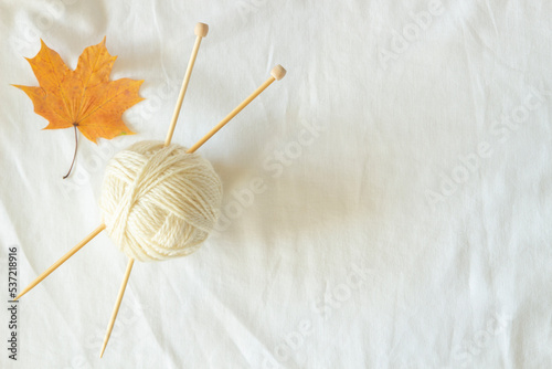 White wool thread ball with wooden bamboo knitting needles and autumn maple fall leaf on white linen fabric background. Top view, copy space. Hobby, relaxation, mental health, sustainable lifestyle