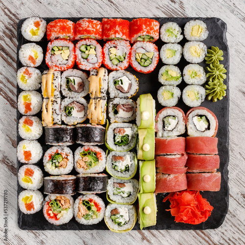 Large assorted variety of sushi rolls served on black metal slate on wooden table.