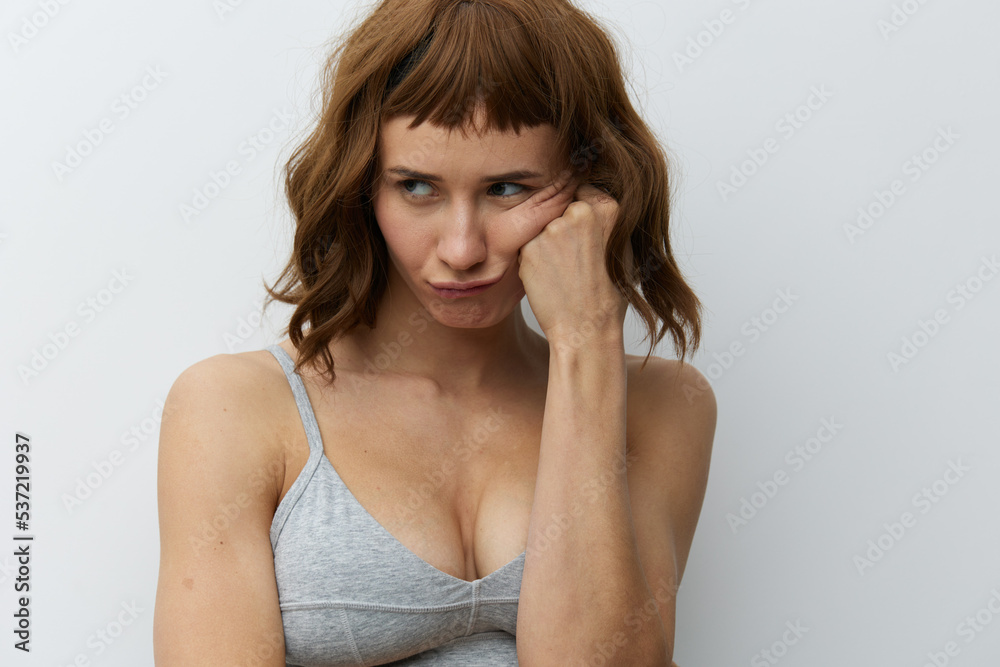 a funny, slightly upset tanned woman with red hair stands on a light background in a gray top and supports her cheek with a fist of one hand a funny, slightly upset tanned woman with red hair stands on a light background in a gray top and supports her cheek with a fist of one hand
