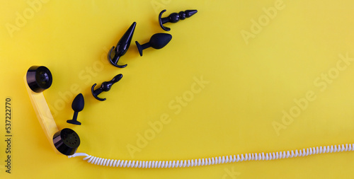 The handset of a yellow retro phone on a yellow background. black sex toys come out of the phone tube. The concept of phone sex, ordering toys for adults. Banner with space for text