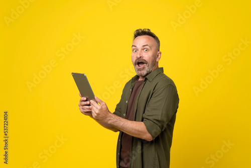 shocked mature man reading news on touchpad. Senior manager wearing formalwear using digital tablet isolated yellow background. Bad news concept. Astonished mustached stubble man with tablet in hands