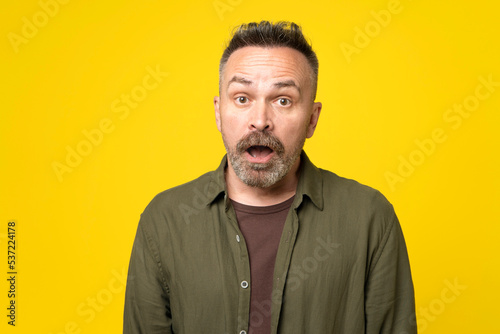 Stubble handsome mature man standing isolated yellow background, looking surprised. Portrait of attractive amazed beard man wearing formal shirt pout lips isolated over bright yellow color background