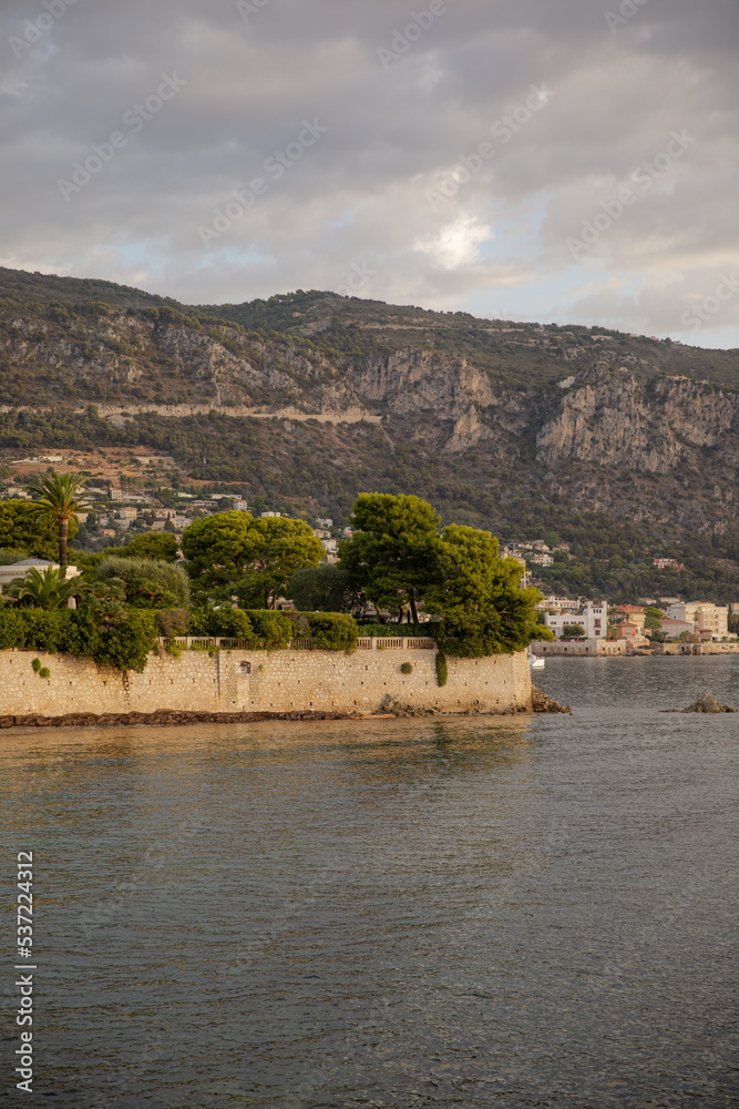 Beautiful view of the small town by the sea. Postcard from Beaulieu-sur-Mer. French coast. Conifer tree against the background of mountains and the sea