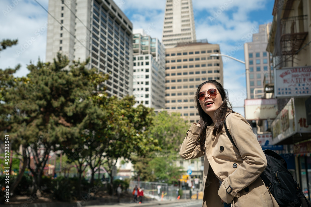 cheerful asian Korean female traveler wearing sunglasses running fingers through hair and looking at nice city landscape while waiting for bus at roadside in san Franciscoâs Chinatown
