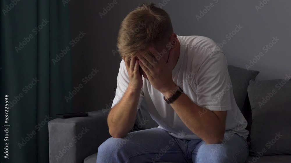 A young man sits on the couch, looking at his phone, touching his face, imitating bad news or disappointment A young man sits on the couch, looking at his phone, touching his face, imitating bad news or disappointment