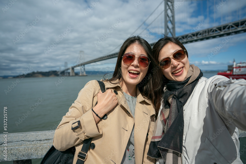 two happy asian Taiwanese girl tourists smiling at camera while taking self portrait together with Oakland bay bridge at background against sunny blue sky in san Francisco California usa