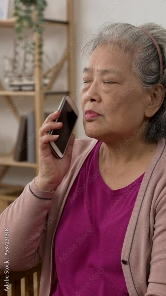 Vertical Screen: asian old lady showing shocked look on face on hearing the surprising news while getting a fraud call on the cell phone in the living room at home