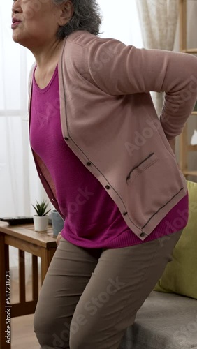 Vertical Screen: chinese elder woman pulling her back muscles as she is standing up from the sofa is groaning painfully and sitting down to rest in the living room at home