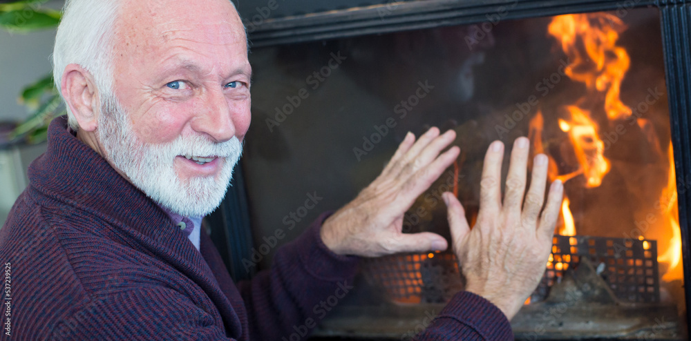 Smiling senior man sitting at fireplace and warming hands in winter 