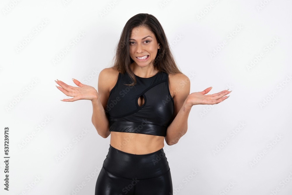 Clueless young beautiful woman wearing sportswear over white background shrugs shoulders with hesitation, faces doubtful situation, spreads palms, Hard decision