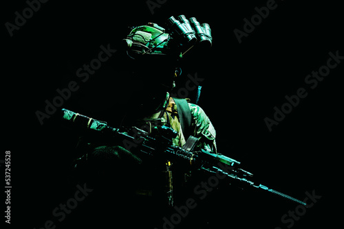 Modern army special forces equipped soldier, anti terrorist squad fighter, elite mercenary armed assault rifle, standing in darkness with night vision goggles on helmet, studio portrait, green light