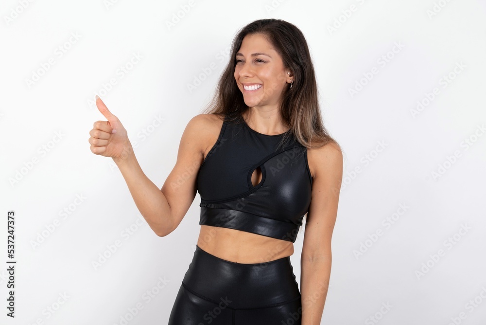 young beautiful woman wearing sportswear over white background Looking proud, smiling doing thumbs up gesture to the side. Good job! young beautiful woman wearing sportswear over white background Looking proud, smiling doing thumbs up gesture to the side. Good job!