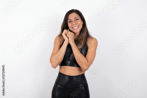 Positive young beautiful woman wearing sportswear over white background smiles happily, glad to receive pleasant news from interlocutor, keeps hands together. People emotions concept.