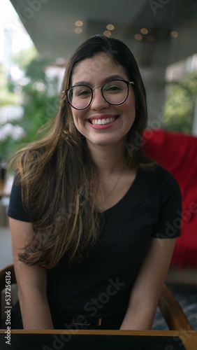 Young woman smiling at camera portrait. Girl in 20s wearing eyeglasses. Person big smile seated at coffee shop in Vertical Video SS