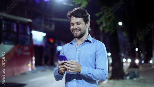 Happy young businessman walking at night in city street holding cellphone smiling. Person commuting after work holding smartphone