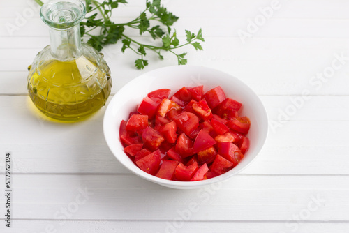 A white bowl with chopped tomatoes, a bunch of fresh parsley and a bottle of olive oil on a white wooden background. Cooking delicious homemade food